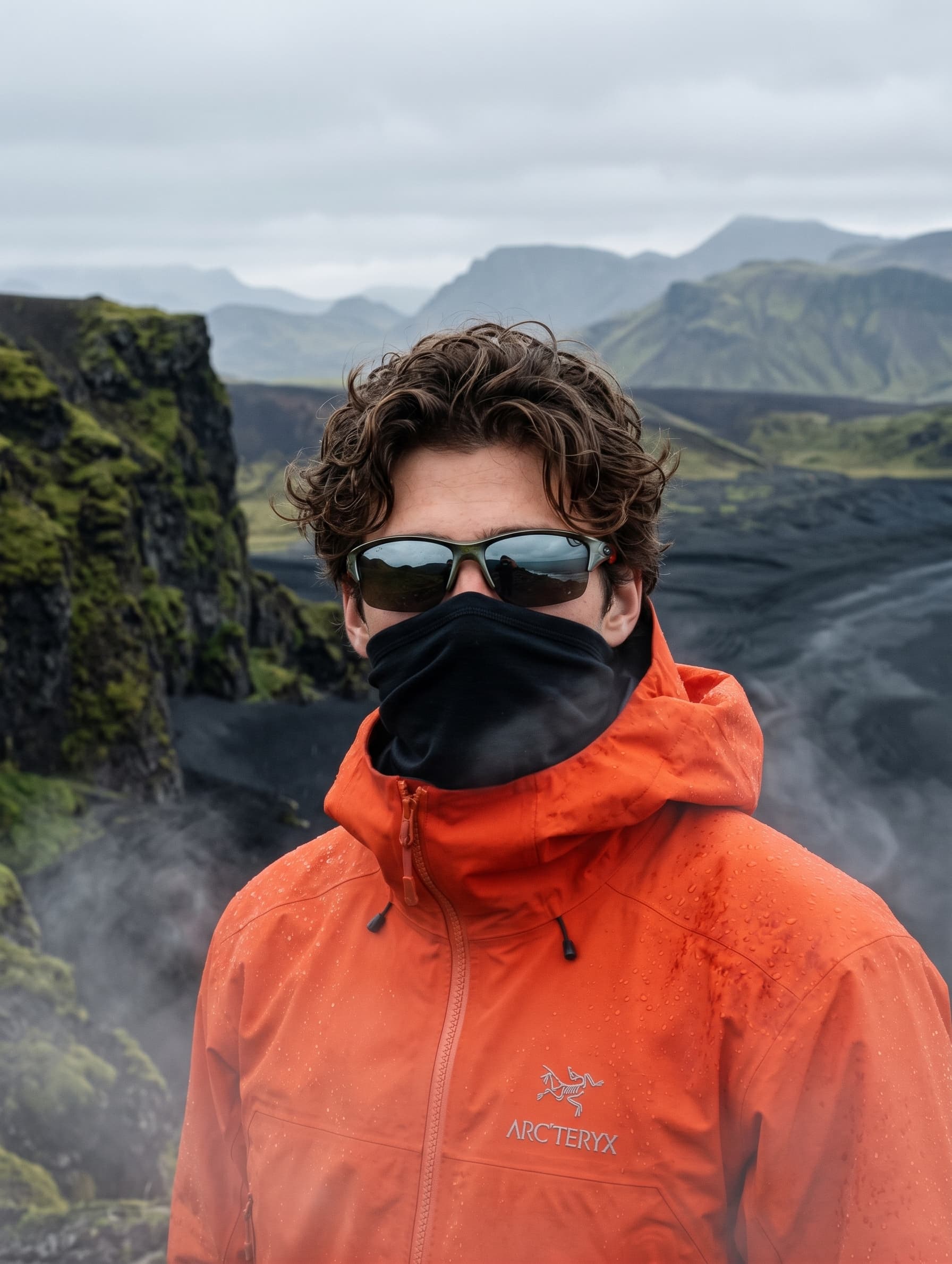 Pessoa de casaco Arc'teryx laranja, óculos escuros e lenço preto, em uma paisagem de serra úmida e nublada
